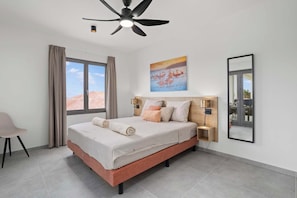 A bedroom with a bed, a ceiling fan, a nightstand, a lamp, a window with curtains, and a painting on the wall. - Bonaire Beach Apartments 6 with a refreshing shared pool and ocean view (Kralendijk)