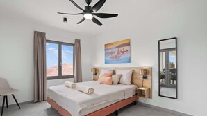 A bedroom with a bed, a ceiling fan, a nightstand, a lamp, a window with curtains, and a painting on the wall. - Bonaire Beach Apartments 6 with a refreshing shared pool and ocean view (Kralendijk)