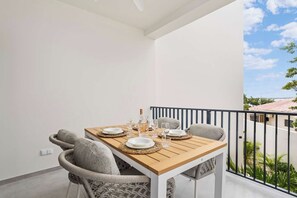 A balcony with a wooden table set for dining, two grey cushioned chairs, and a view of a building and greenery.