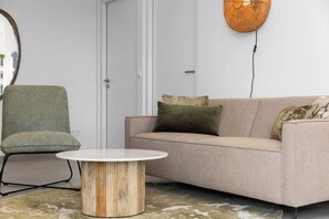 Un salon avec un fauteuil vert, un canapé beige orné de coussins à motifs et une table basse ronde en bois.