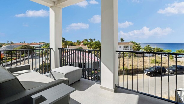 Een balkon met uitzicht op de oceaan, een terras met tuinmeubilair en een parkeerplaats met auto's.