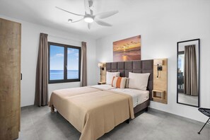 Un dormitorio con una cama grande, un ventilador de techo, una ventana con vista al mar y un cuadro de un atardecer.
