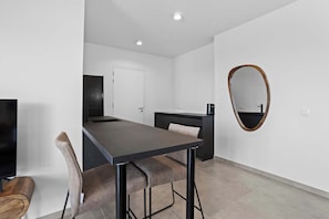 A modern, minimalist room with a dark wooden dining table, two upholstered chairs, a flat-screen TV, a small kitchenette, and a large mirror on the wall.