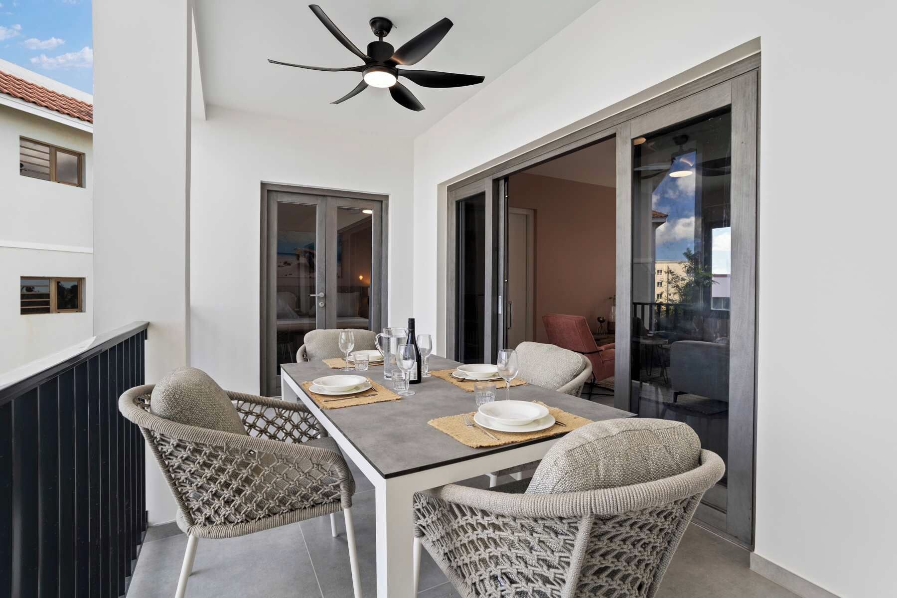 A balcony with a dining table set for two, featuring wicker chairs and a ceiling fan.