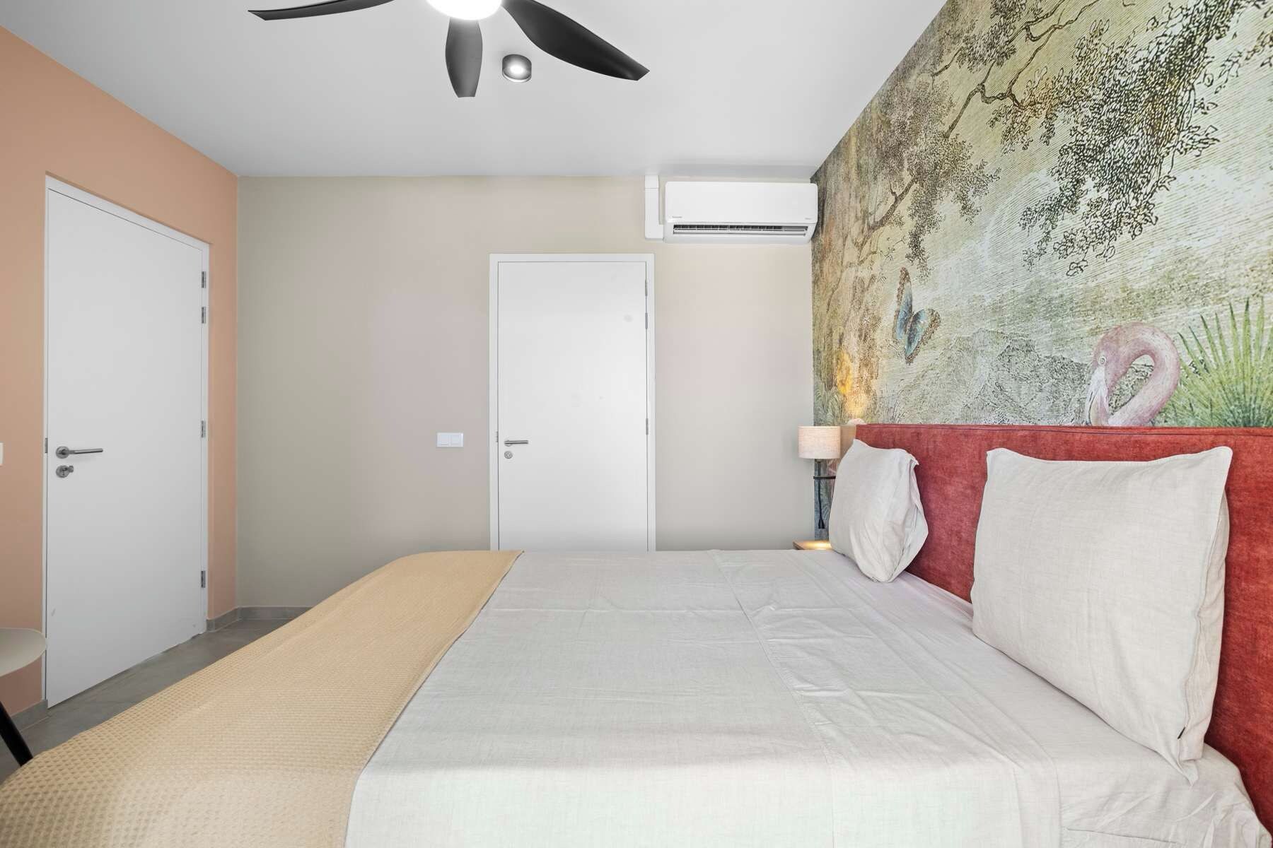 A bedroom with a bed, a ceiling fan, an air conditioning unit, a door, and a wall with a painting.