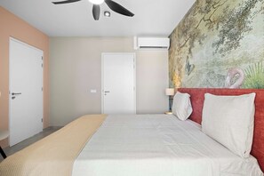 A bedroom with a bed, a ceiling fan, an air conditioning unit, a door, and a wall with a painting.