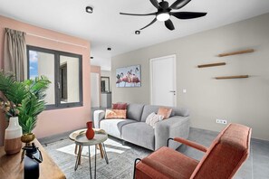 A modern living room with a grey sofa, a red armchair, a round coffee table, and a ceiling fan. There is a large window with curtains, a potted plant, and a painting of flamingos on the wall.