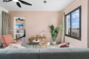 Un salon moderne avec un canapé gris, une table basse en bois, une petite table d’appoint, une plante en pot et une vue sur le ciel par une grande fenêtre.