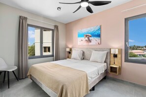 Une chambre à coucher comprenant un lit, deux tables de chevet, un ventilateur de plafond, une grande fenêtre et un tableau représentant une tortue de mer.
