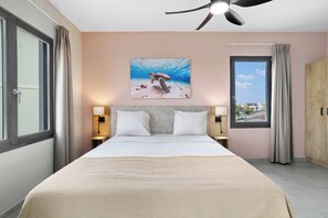 Habitación de hotel con cama, dos lámparas de noche, ventilador de techo, ventana grande con cortinas y un cuadro de tortuga marina en la pared.