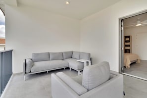 A modern, minimalist living room with a grey sectional sofa, a small white side table, and a glass door leading to a bedroom.