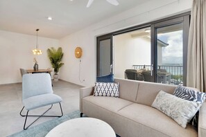 Ein modernes Wohnzimmer mit einer Couch, einem Sessel und einem runden Couchtisch. Es gibt einen Essbereich mit Tisch und Stühlen sowie einen Balkon mit Blick auf das Wasser.