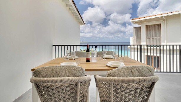 A balcony with a wooden table set for two, two wicker chairs, a bottle of wine, and a glass of water. - Bonaire Beach Apartments 15 with a refreshing shared pool and spectacular sea view (Kralendijk)