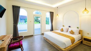 A bedroom with a large bed, a wooden nightstand, a desk with a chair, and a view through the window. - Mahayana House (Tabanan)