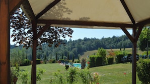 A covered outdoor area with a table and chairs, surrounded by greenery and a view of a pool and hills. - Charming Flat With Pool Surrounded by Nature (Asciano)
