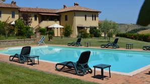 A pool area with lounge chairs and umbrellas, surrounded by a landscaped garden and a building with a tiled roof. - Charming Flat With Pool Surrounded by Nature (Asciano)