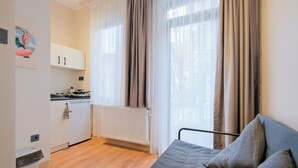 Un espace de vie compact comprenant un coin cuisine, un lit avec une literie grise et une fenêtre avec des rideaux.
