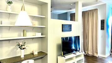 A modern living room with a flat-screen TV on a white stand, a white pendant light, and a shelf with decorative items.