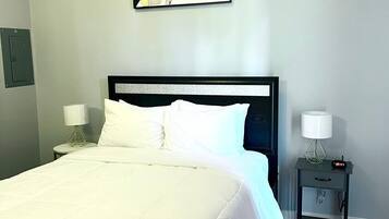 A neatly made bed with white bedding, a black headboard, and a painting above it. There is a small bedside table with a lamp and a clock.