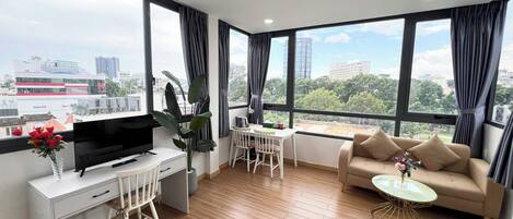 A modern living room with a TV, desk, and sofa, large windows with curtains, and a city view.