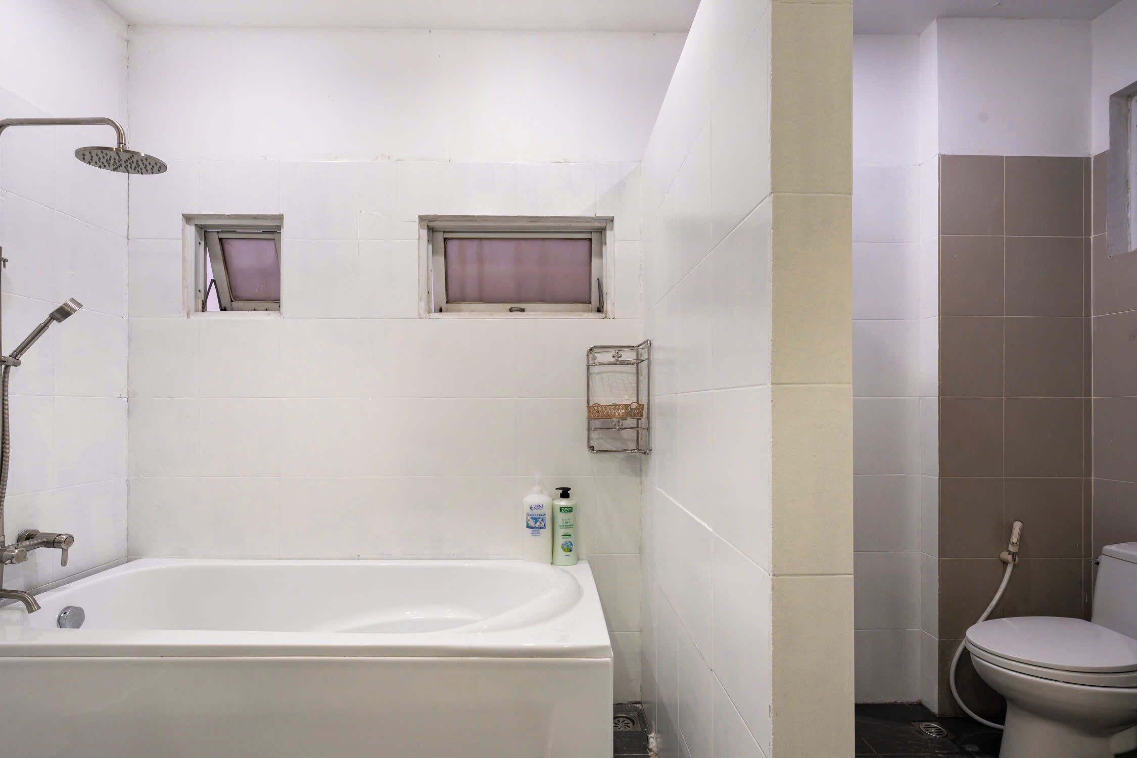 Een badkamer met een bad, toilet en douche. Er zijn twee kleine ramen en een zeepdispenser op de muur.