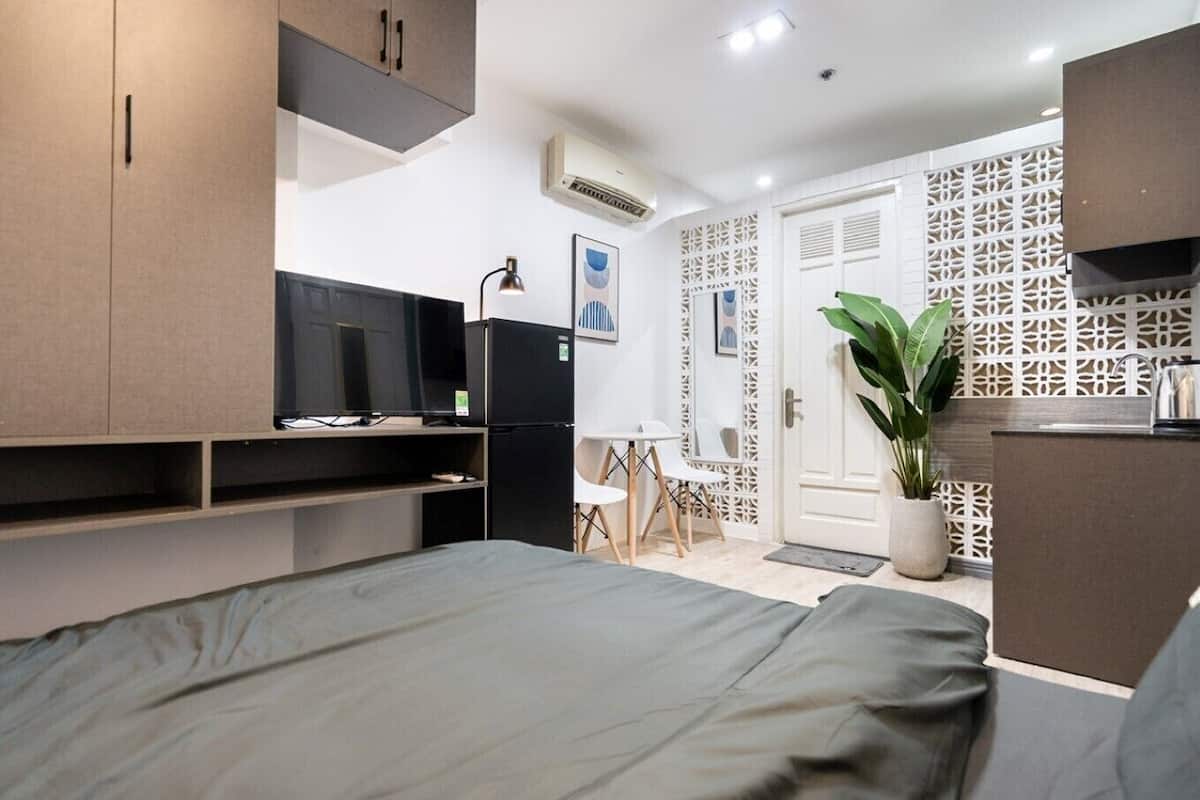 A modern hotel room with a bed, a desk with a computer, a TV, and a refrigerator. There is a potted plant and a decorative wall panel.