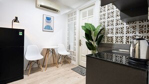Cocina compacta con un refrigerador negro, una mesa blanca con dos sillas, una planta en maceta y un hervidor de acero inoxidable.