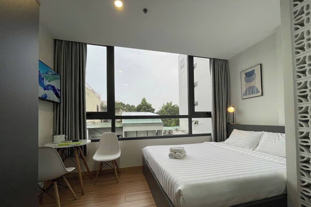 A hotel room with a bed, a desk with a chair, a window with a view of buildings, and a wall with a decorative panel.