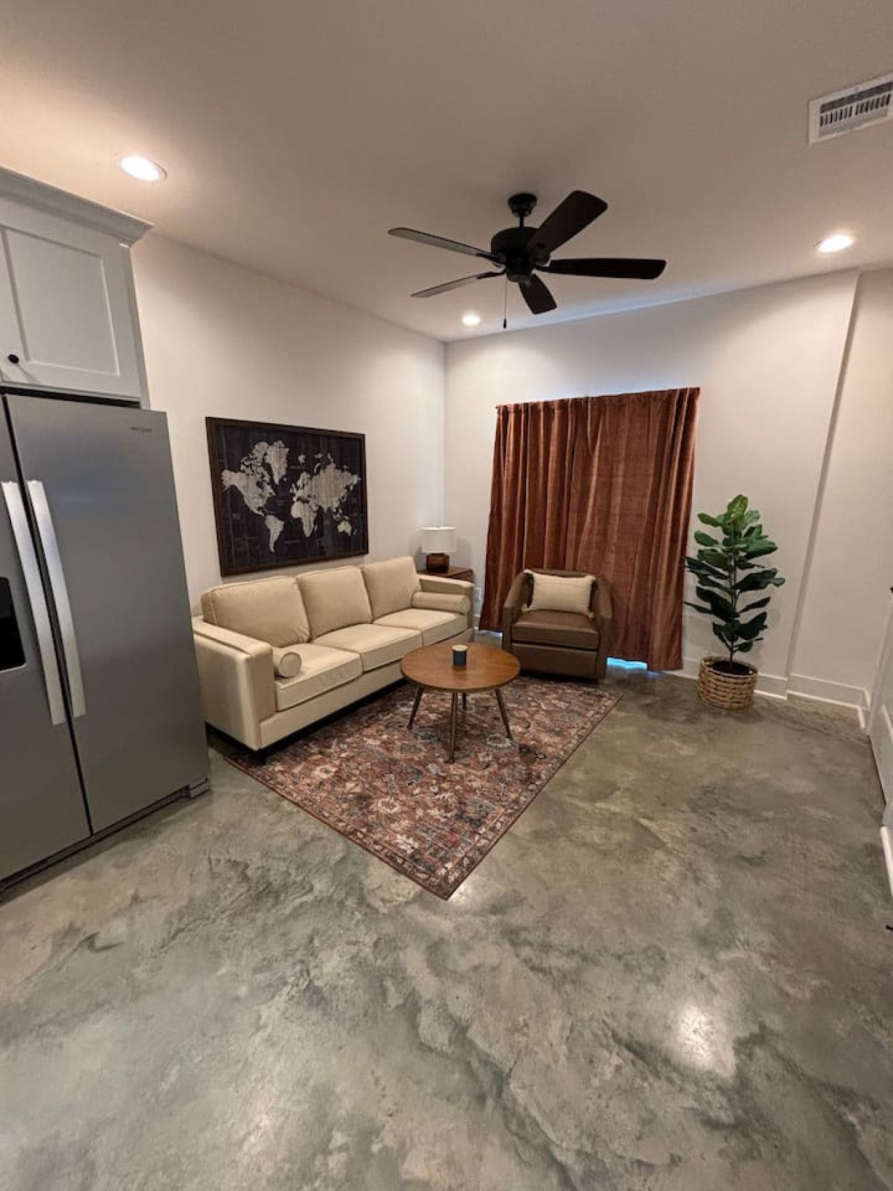 Un salon moderne avec un canapé beige, une table basse, un lampadaire, une œuvre d’art représentant une carte du monde, un ventilateur de plafond et une plante en pot.