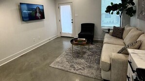 A spacious living room with a high ceiling, a ceiling fan, a flat-screen TV mounted on the wall, a beige couch, a black armchair, and a coffee table. - NEW-Beignet House-Luxe Mattress-Fast WiFi-Book Now (Lafayette)