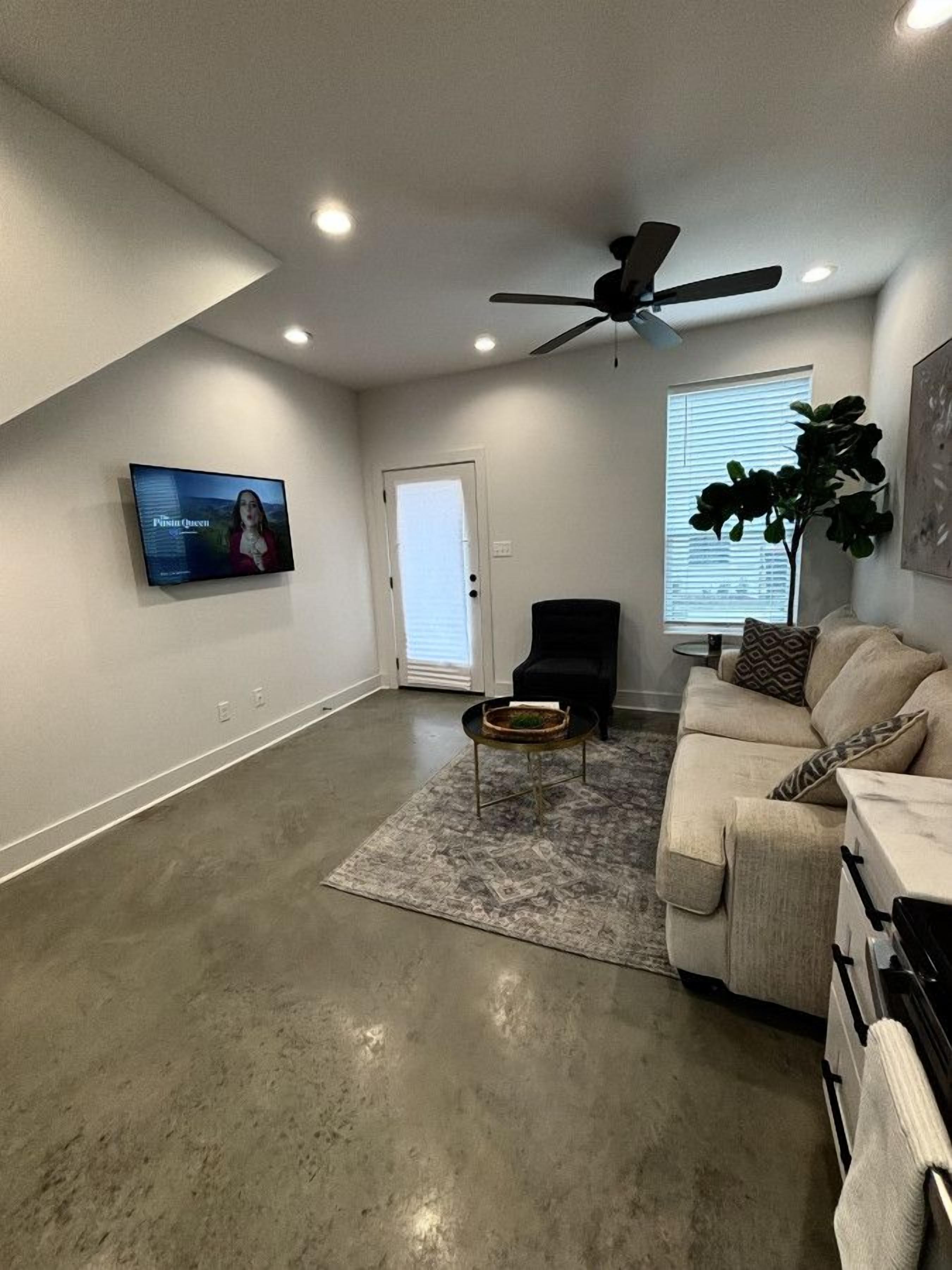 A spacious living room with a high ceiling, a ceiling fan, a flat-screen TV mounted on the wall, a beige couch, a black armchair, and a coffee table.