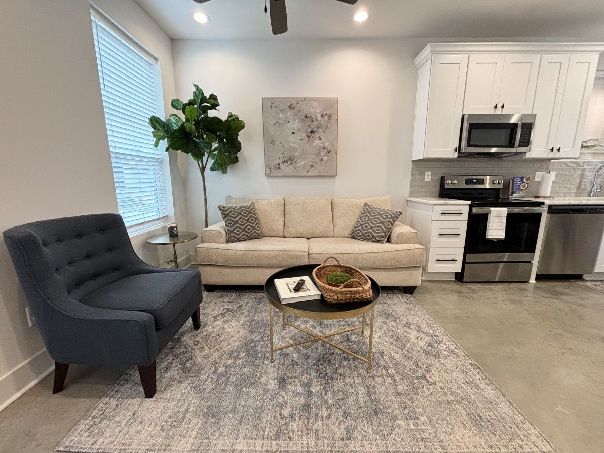 Un salon moderne avec un fauteuil foncé, un canapé beige et une table basse sur laquelle se trouve un panier. Une grande plante est placée à côté du canapé, et un tableau est accroché au mur.