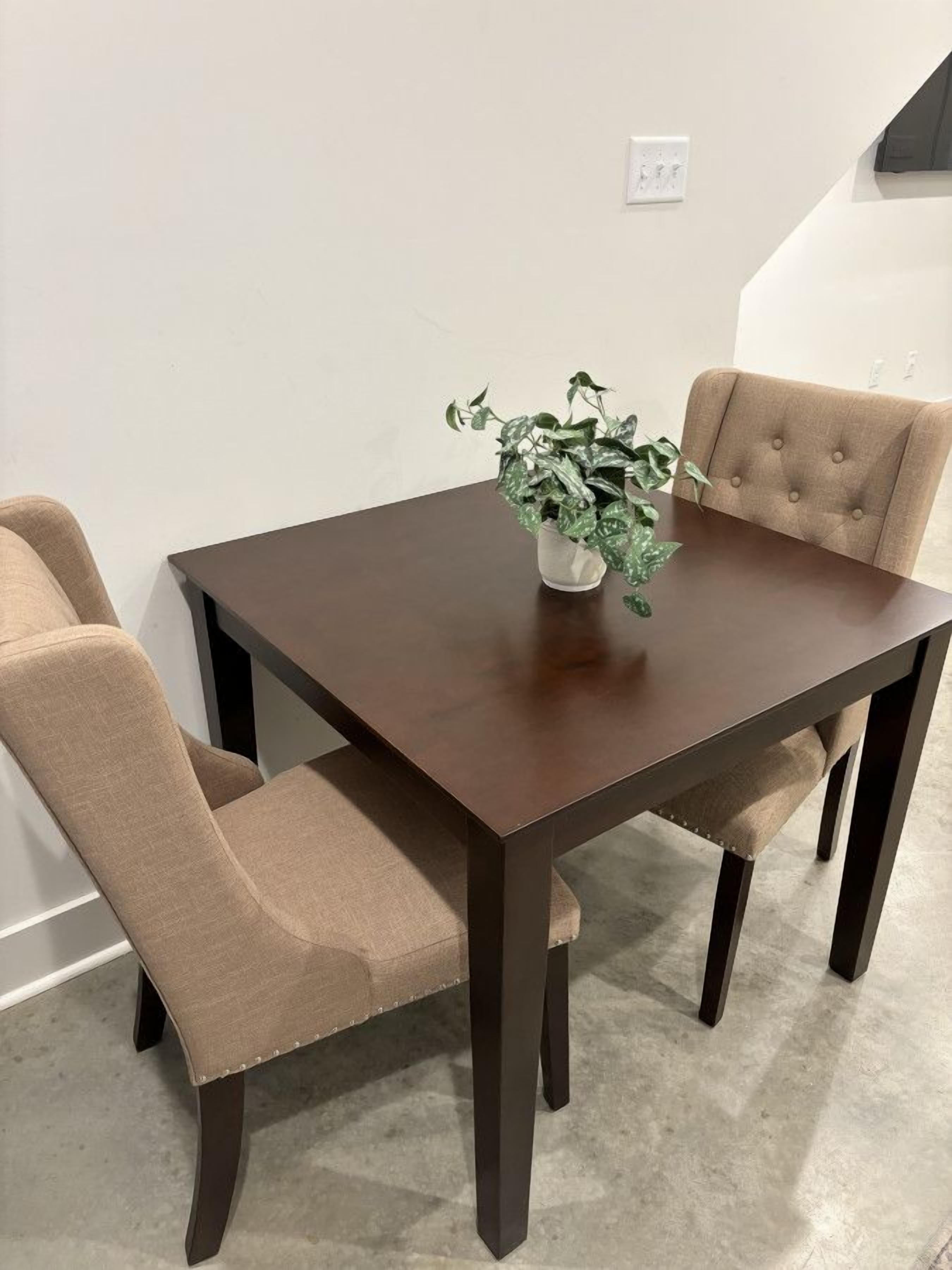 A dining table with four beige upholstered chairs, a potted plant on the table, and a white wall with a power outlet in the background.