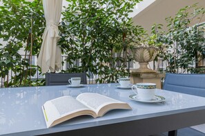 Mesa al aire libre con un libro, dos tazas y platillos, rodeada de follaje y una fuente de piedra.