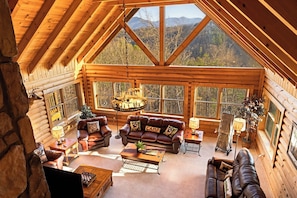 L’intérieur d’une cabane en rondins, avec une chambre à coucher située en mezzanine, des murs en bois, une cheminée et une grande fenêtre donnant sur une forêt.