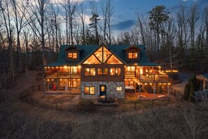 A large log cabin with multiple decks and a stone fireplace, surrounded by a forest.