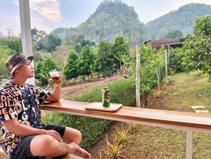 A person sitting at a wooden table with a drink and a can of beer, overlooking a lush green landscape with trees and a hill in the background. - GJ TOUR and HOME STAY (Pangmapha)