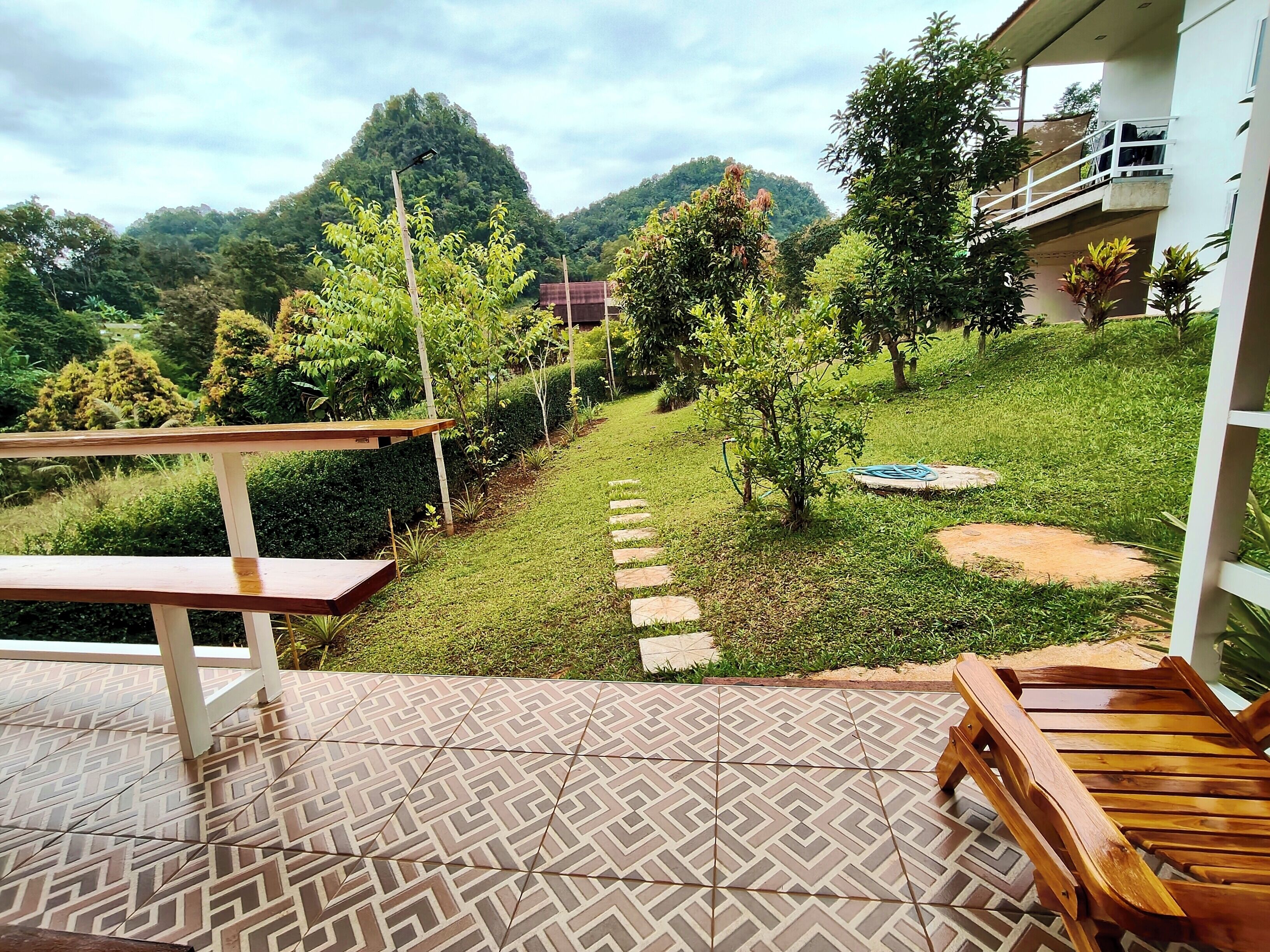 A patio with patterned tiles, a wooden bench, and a view of a lush garden with a pathway leading to a building.