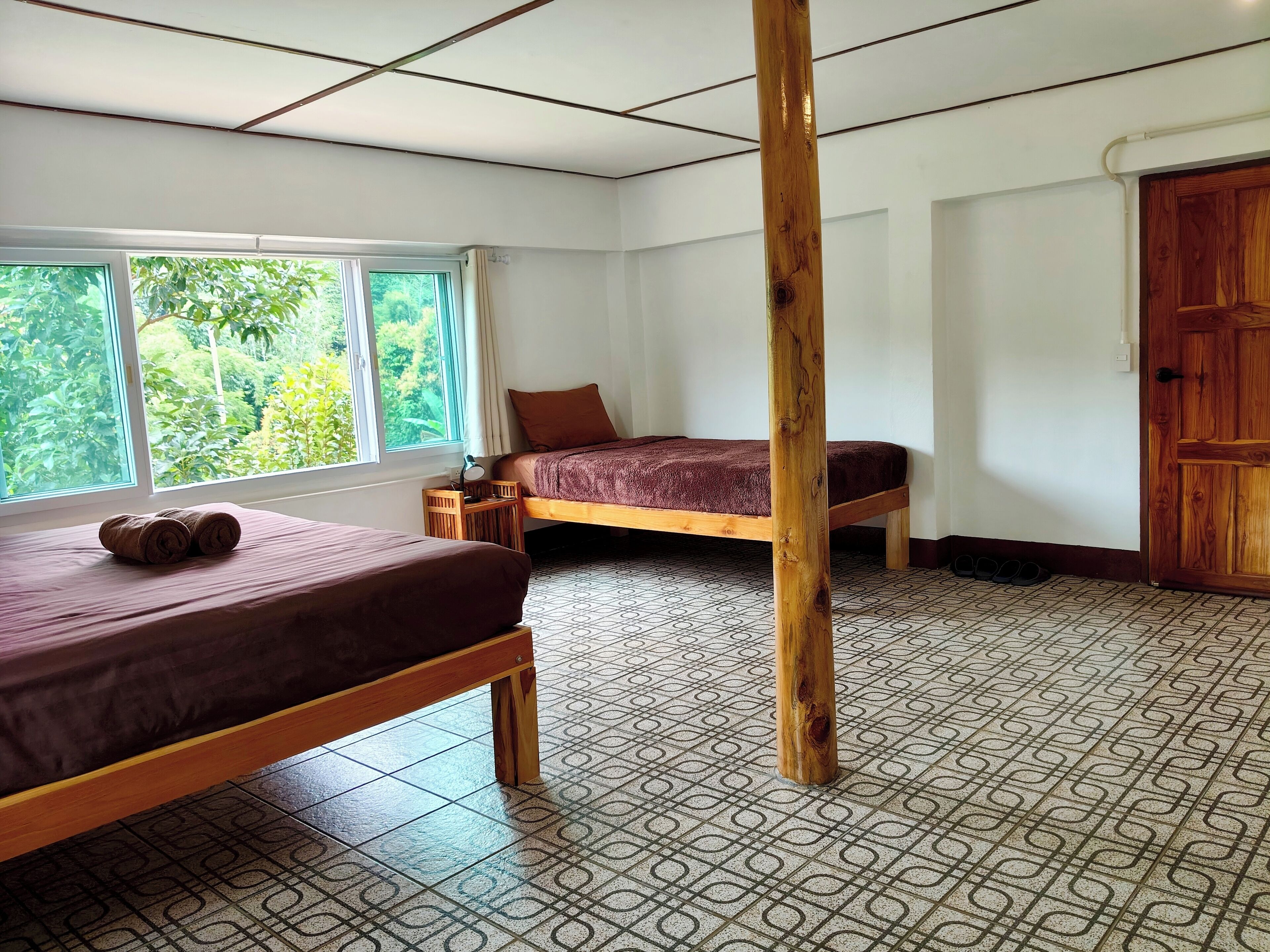 A room with two beds, a wooden pillar, a window with a view of greenery, and patterned floor tiles.