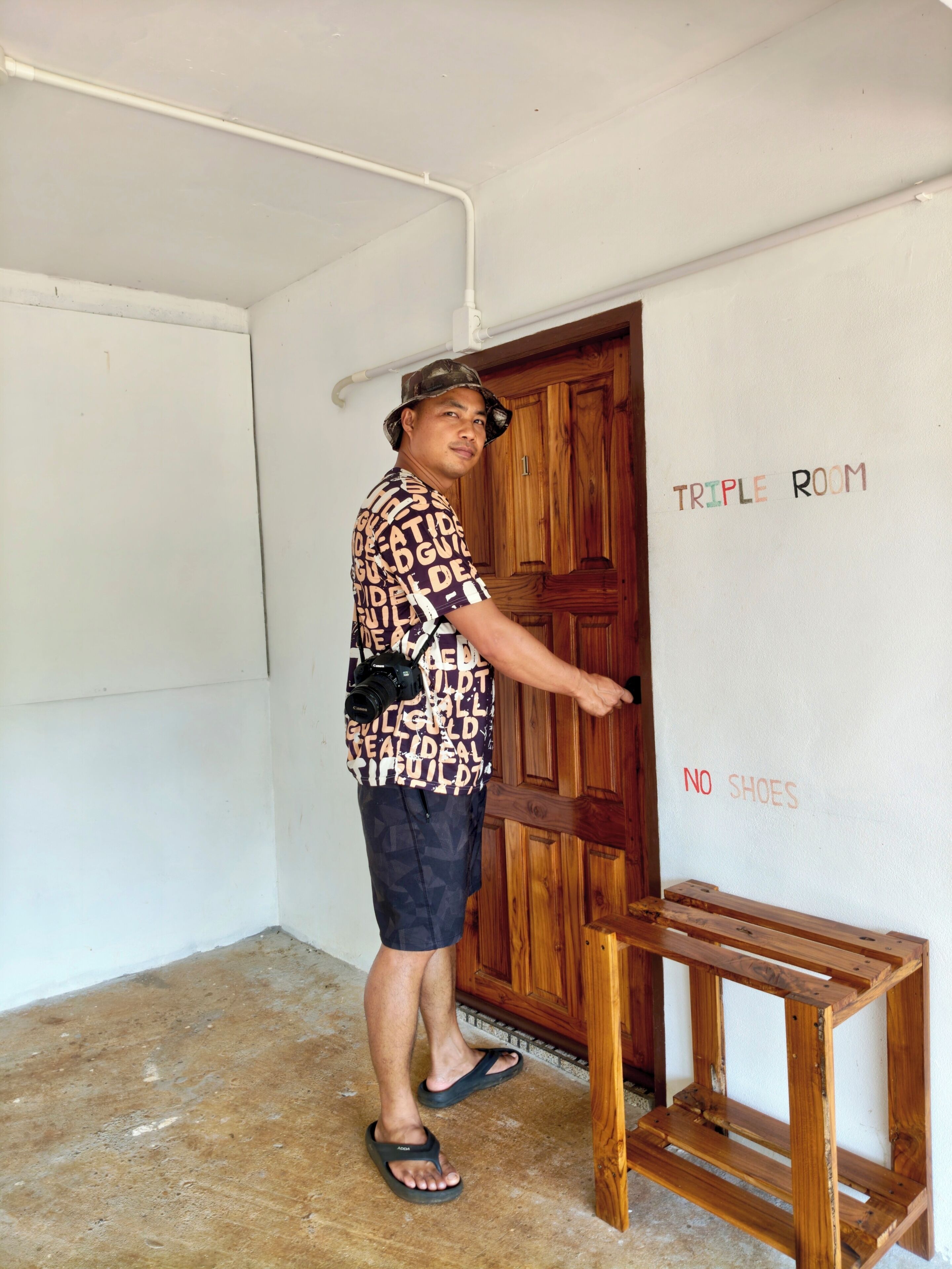 Una persona de pie en una habitación con una puerta de madera, una pequeña mesa de madera y una pared con la leyenda 'TRIPLE ROOM' y 'NO SHOES'.