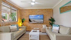 A living room with brick walls, a ceiling fan, and a beach view painting. There are two sofas with matching pillows and a small white table. - Jervis Joy – child friendly coastal retreat (Callala Beach)