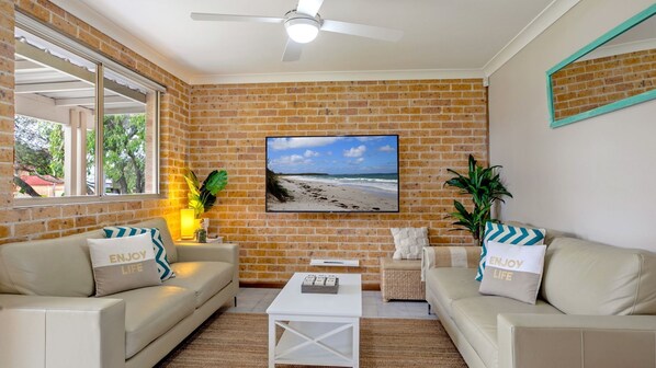 A living room with brick walls, a ceiling fan, and a beach view painting. There are two sofas with matching pillows and a small white table.
