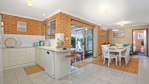 A modern kitchen with a brick wall, a dining area with white furniture, and a sliding glass door leading to a balcony. - Jervis Joy – child friendly coastal retreat (Callala Beach)