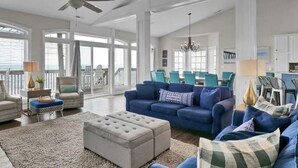 A spacious living room with a large window, a blue sofa, a beige ottoman, and a white armchair. - Surfer's Delight - Amazing 6 BR Oceanfront Home in Hampton Colony (North Topsail Beach)