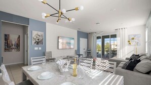 A modern dining area with a marble table, white chairs, and a chandelier. There is a sofa and a dining table with chairs in the background.