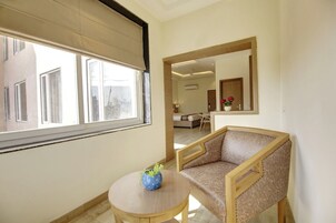 A room with a large window, a round table with a vase, a sofa, and a mirror reflecting the room.
