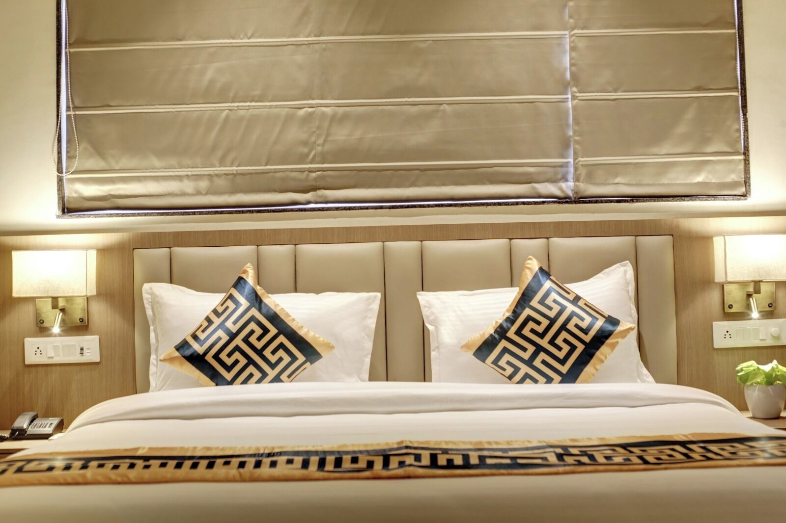 A hotel room with a large bed, two decorative pillows, a nightstand with a lamp, and a window with blinds.