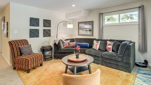 Een woonkamer met een gestreepte fauteuil, een bank met kleurrijke kussens, een ronde salontafel met een bloevas en een groot raam met gordijnen.