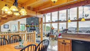 A wooden interior with a dining area, kitchen, and large windows offering views of the outdoors. - Hot Tub + Games! Mtn-view Cabin Near Pigeon Forge (Sevierville)