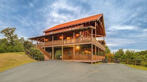 A two-story log cabin with a balcony and a porch, surrounded by a grassy area and trees. - Hot Tub + Games! Mtn-view Cabin Near Pigeon Forge (Sevierville)